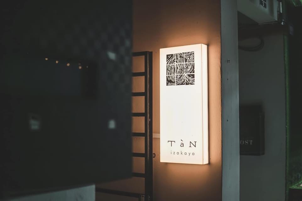 The lit TàN izakaya signboard on the wall at the entrance